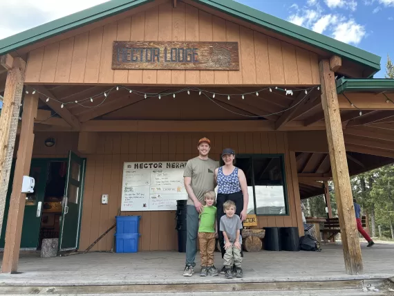 A family smiles for a photo in front of Hector Lodge at Camp Chief Hector YMCA