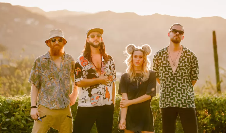 Walk Off the Eart, a band, stands in front of a mountain backdrop at golden hour, with 3 of the 4 members wearing sunglasses