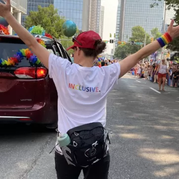 Individual with arms raised in the shape of a y with a white t shirt on that says inclusive on the back